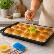 A person brushes golden-brown baked buns with a green silicone brush in a black baking pan on a white marble counter.