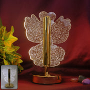 A glowing crystal butterfly lamp with a gold stem and base, against a dark red background with green foliage and small rocks.