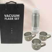 A black gift bag and a silver vacuum flask with three beige mugs are displayed against a softly blurred background.