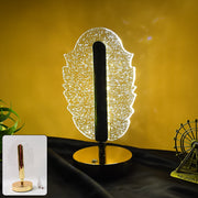 A glowing feather-shaped crystal table lamp with a gold base and a small inset of the lamp on a dark surface.