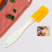 A yellow silicone spatula with a cream handle lies on a grey surface, next to a red fabric and a small inset of a hand using