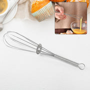 A silver spring coil whisk is displayed on a white surface with a blurred background of baked goods and person whisking