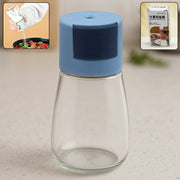A clear glass spice dispenser with a blue top stands on a kitchen counter, with an inset of it in use.