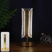 A lit gold and clear crystal table lamp on a dark wooden surface, with a small cannon and plant in the background.