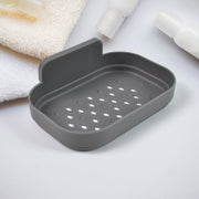 A gray self-adhesive soap dish with drainage holes is shown on a white surface with towels and bottles.