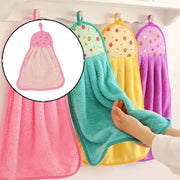 A hand holds a teal microfiber towel with a fruit print top, hanging among other colorful towels on hooks in a kitchen.
