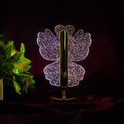 A glowing crystal butterfly-shaped table lamp with a gold base and stem, against a dark background with green foliage.