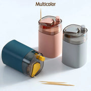 Three automatic toothpick dispensers in teal, pink, and grey, arranged on a white surface with two toothpicks in the
