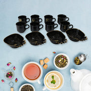 A black ceramic tea set with eight cups and four pineapple-shaped snack bowls on a blue surface, surrounded by tea leaves