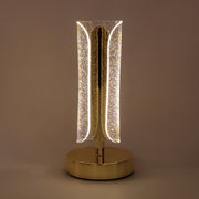 A lit gold and clear crystal table lamp with a curved design stands alone on a plain background.