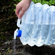 A hand holds a translucent collapsible water container with a blue spigot, pouring water onto the ground in a natural