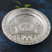 A round silver-colored plastic pooja thali with intricate embossed floral and geometric patterns, with green leaves in the