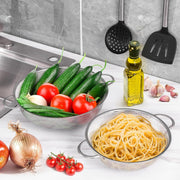 Two stainless steel strainer bowls, one with cucumbers and tomatoes, the other with pasta, on a kitchen counter.