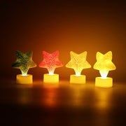 Four glowing star-shaped LED tealight candles in green, red, and two yellow, arranged in a row in a dark setting.