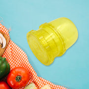 A yellow plastic manual food grinder is angled on a blue background, with a red and white checkered cloth and vegetables in