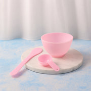 A pink mixing bowl, spatula, and measuring spoon are arranged on a round marble tray against a white curtain.