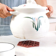 Hands hold a large white ceramic pot with green and red floral designs over a single maroon silicone coaster.