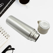 The silver vacuum flask lies on a white desk next to its detached beige cap, with a notebook and glasses nearby.