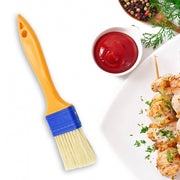 A yellow and blue basting brush is positioned next to a bowl of red sauce and skewers of grilled chicken.