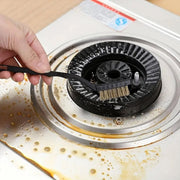 A hand uses a black-handled brass wire brush to clean a very dirty gas stove burner.