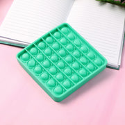 A square green silicone pop-it fidget toy resting on a pink surface next to an open notebook.