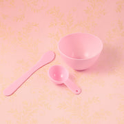 A pink mixing bowl, spatula, and measuring spoon are arranged on a pink background with a subtle floral pattern.