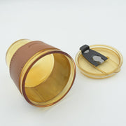 An amber glass cup with a brown leather sleeve lies on its side next to its matching amber lid on a white surface.