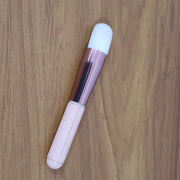 A pink and rose gold makeup brush with a white brush head, angled on a wooden surface.