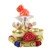A white Ganesha idol with an orange turban sits on a gold base with two empty gold bowls and a red rose, against a white