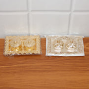 Two ornate rectangular pooja thalis, one gold and one silver, both in clear plastic packaging, on a wooden surface.