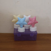 Four unlit star-shaped LED tealight candles in yellow, blue, white, and pink, displayed on a purple plastic box.