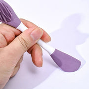 A hand holding the purple and white double-headed silicone mask brush, showing both ends.