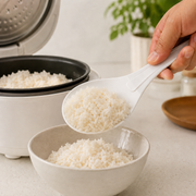 A hand uses a white rice paddle to scoop white rice from a rice cooker into a bowl.
