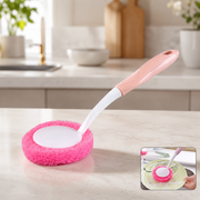 A pink and white long-handled dish scrubber with a round pink scrubbing pad rests on a kitchen counter.