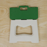 A green and white plastic folding stool is shown partially folded, revealing its carry handle, on a wooden surface.