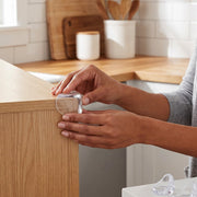 A person's hands applying a clear, rounded corner protector to a light wood table edge in a kitchen.