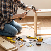A person in a plaid shirt and jeans kneels on a wooden floor, using a hammer to assemble a light wood frame.