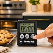 A hand presses the 'STOP START' button on a black digital timer displaying 05:00, next to a tray of cookies.