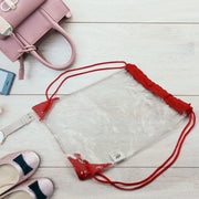 Clear drawstring bag with red cords and accents, surrounded by a pink handbag, sparkly shoes, and a watch on a wooden floor.
