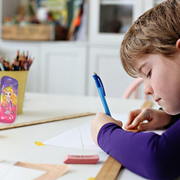 A child drawing with a blue pen and ruler, with a mermaid pencil case standing upright in the blurred background.
