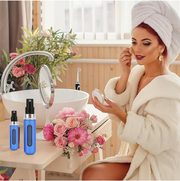 A woman in a white robe and towel applies makeup in a bathroom, with two blue refillable perfume bottles on a wooden counter