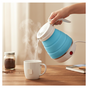 A hand pours hot water from a blue and white foldable electric kettle into a white mug with a tea bag on a wooden table.