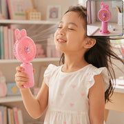 A young girl with closed eyes smiles as she holds a pink bunny ear mini handheld fan to her face, feeling the breeze