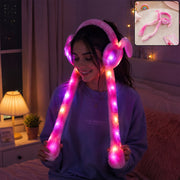 A young woman smiling while wearing pink LED light-up plush bunny earmuffs with long tails that glow brightly in dimly 