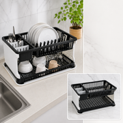 A black and white two-tier dish drying rack with dishes and utensils on a kitchen counter next to a sink.