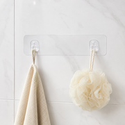 A clear adhesive wall hook strip with two hooks, holding a beige towel and a cream bath pouf against a white tiled wall.