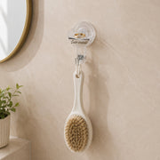 A clear self-adhesive hook holds a white-handled body brush with natural bristles on a light tiled bathroom wall next to a