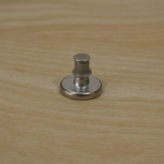 A silver metal button pin with its back lock attached, sitting on a light wooden surface.