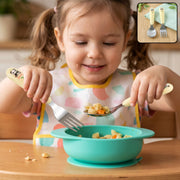 A young girl in a bib holds a panda-handled fork and spoon, eating pasta from a teal bowl.