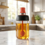 A glass dispenser filled with honey, featuring a red silicone dipper and black lid, on a kitchen counter.